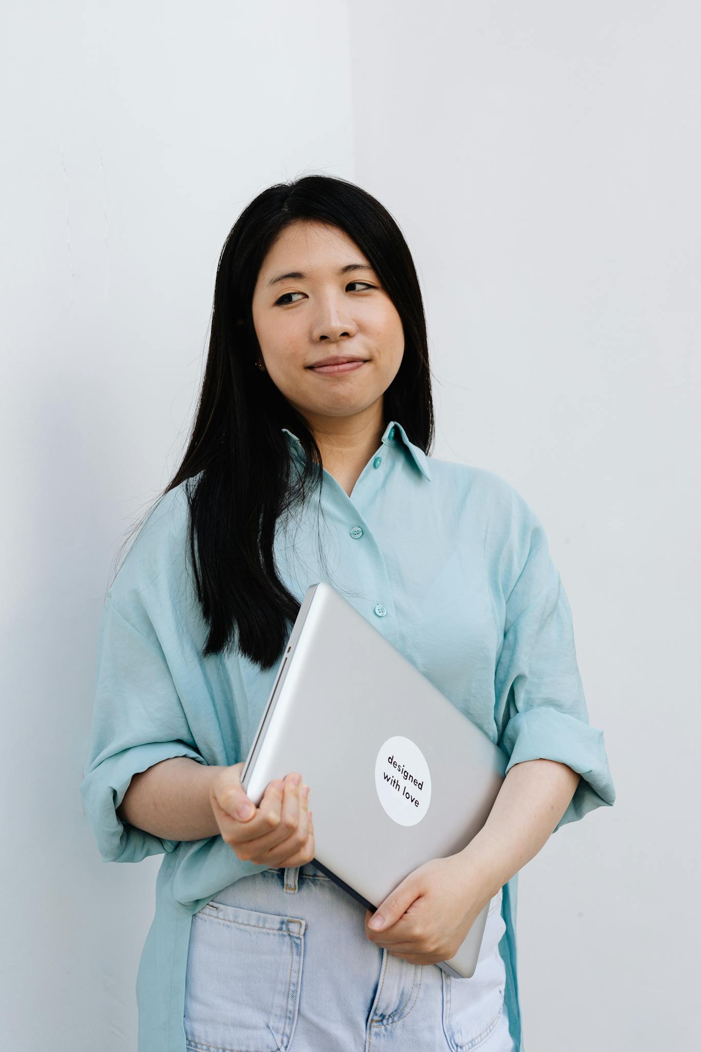 Asian woman in casual attire holding a laptop indoors against a white background.