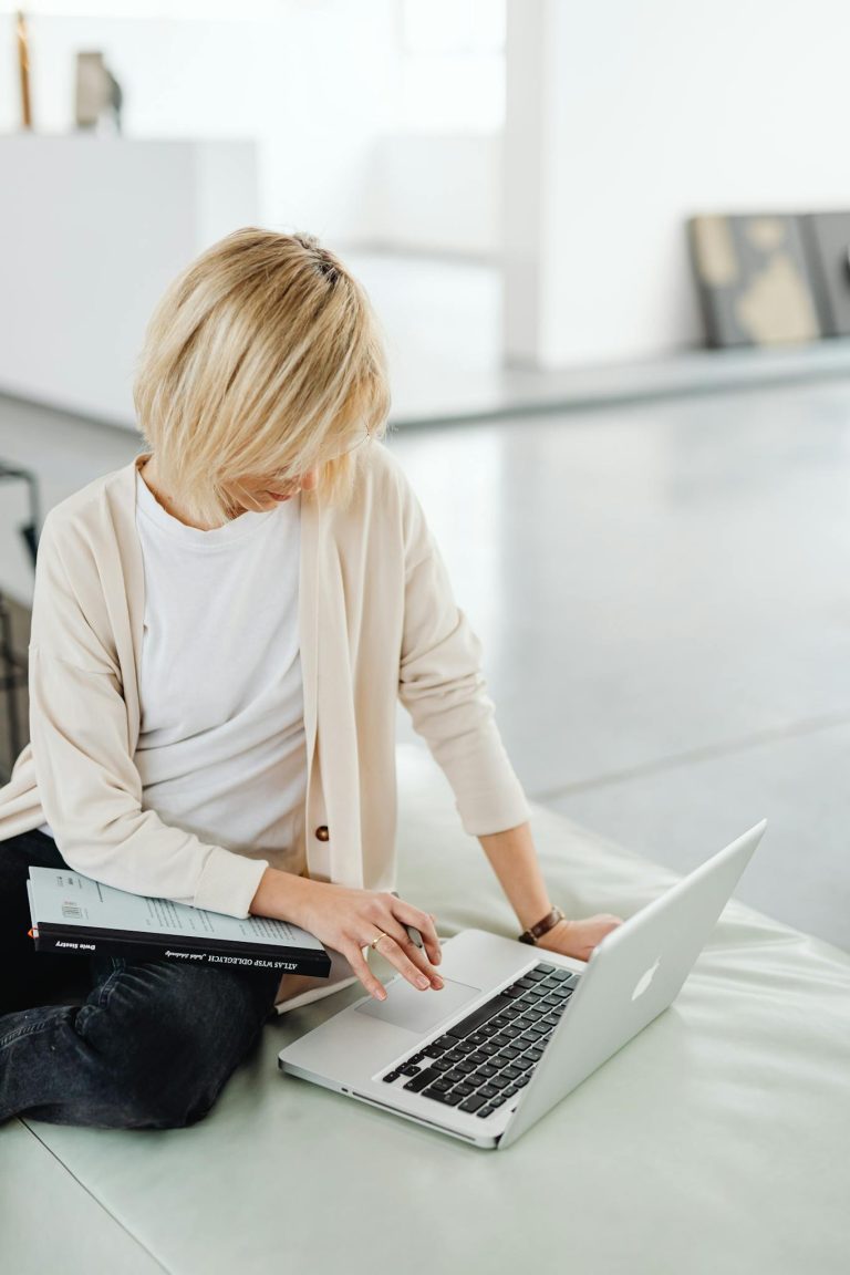 Blonde woman sitting casually using a laptop indoors on a bright day.