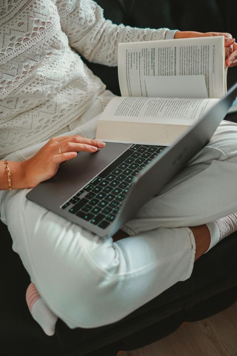 Woman sitting cross-legged with laptop and book, focused on studying indoors.