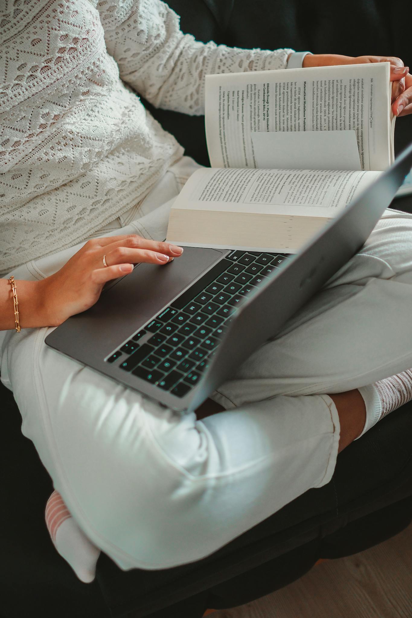 Woman sitting cross-legged with laptop and book, focused on studying indoors.