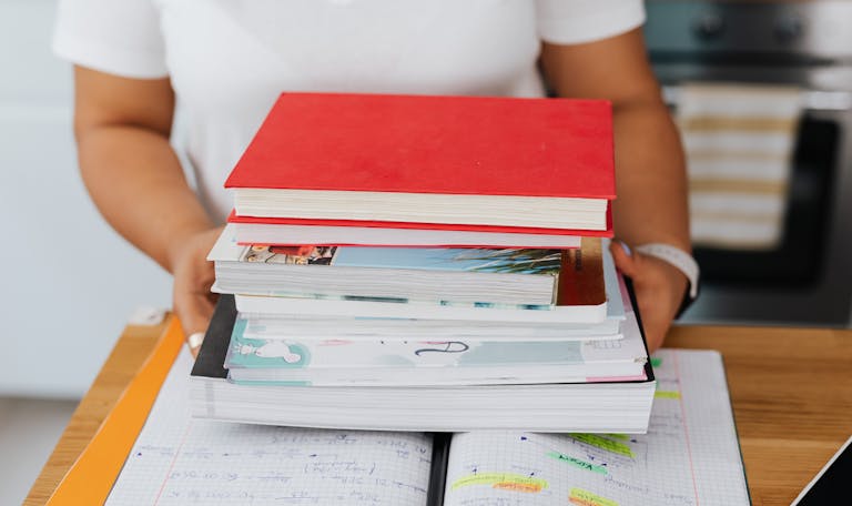 Close-up of a person holding a stack of colorful books over an open notebook.