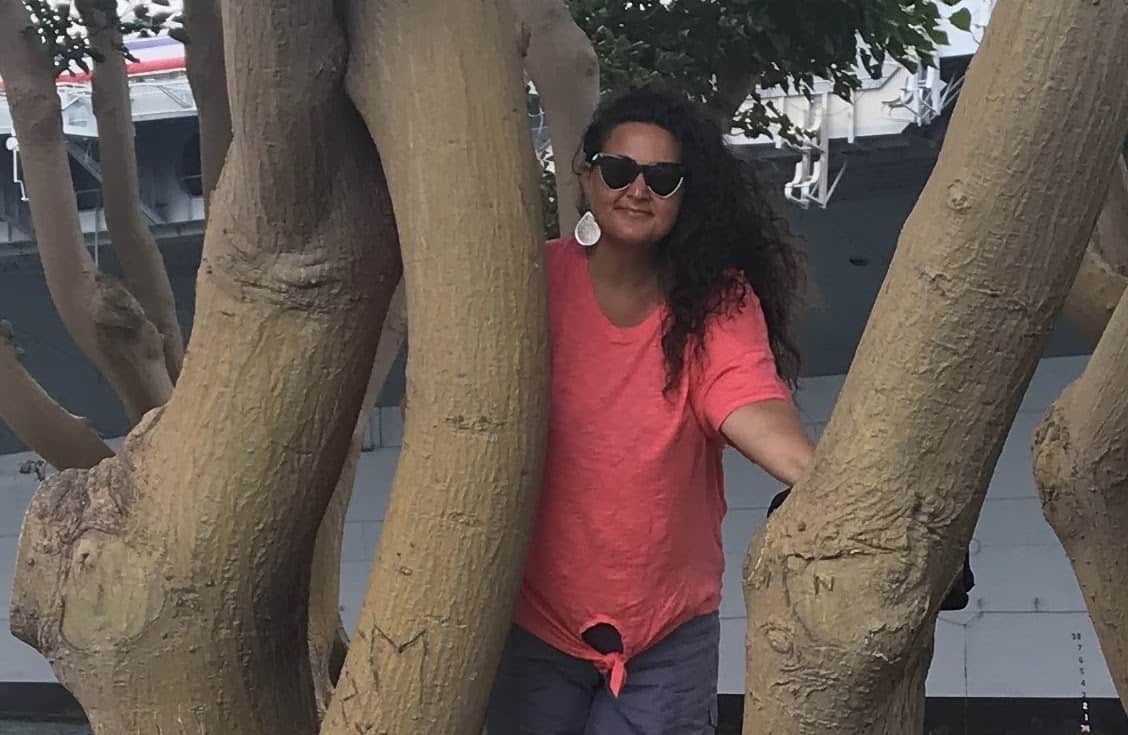 Woman wearing sunglasses and a bright pink shirt leaning up against a tree