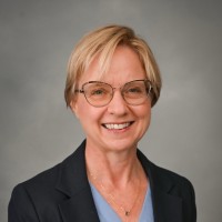 person with short blond hair wearing glasses, a blue shirt, and black blazer